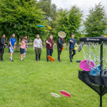 A group of people lined up on grass in a park, enthusiastically throwing colourful discs toward a disc golf basket in the foreground. Several discs are mid-air, flying toward the target, with some already landing in or near the basket. The participants are smiling and engaged, surrounded by green trees. The image captures the fun, energy, and inclusivity of an Active Cities Dublin outdoor event.